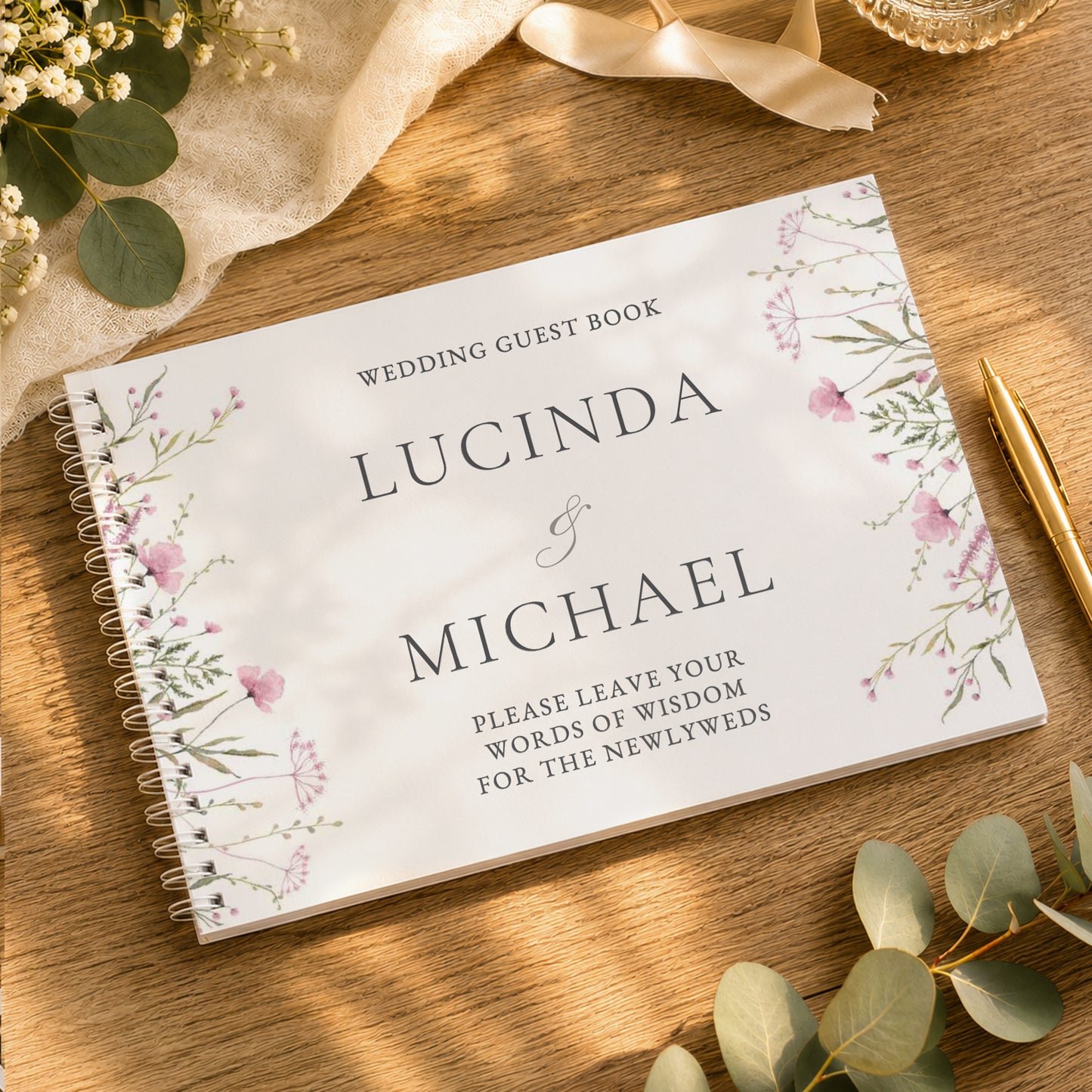 A personalised spiral-bound wedding guest book titled 'Lucinda & Michael' with delicate pink and green floral illustrations, styled on a wooden table with eucalyptus leaves, a gold pen, and a cream ribbon.