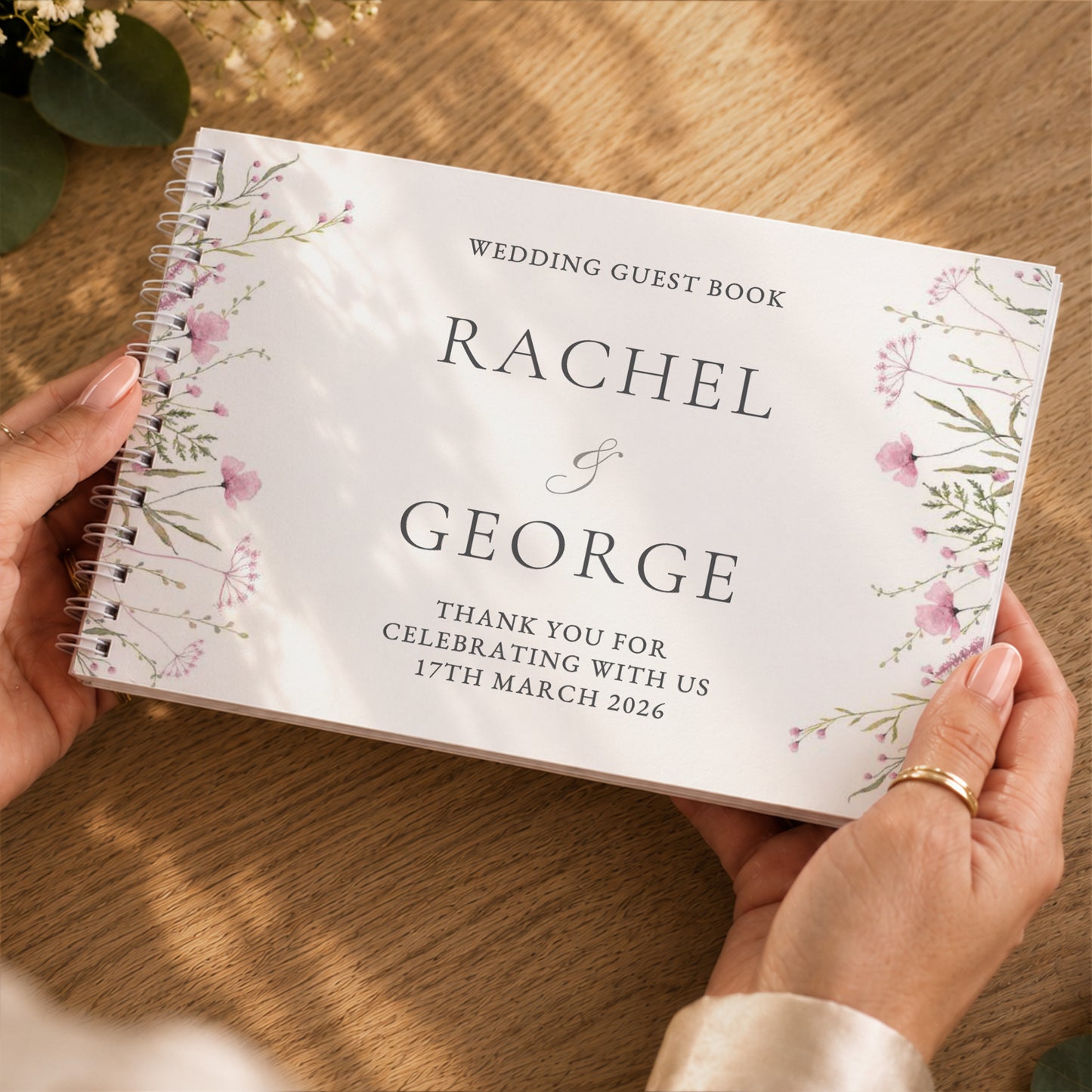 A personalised spiral-bound wedding guest book held by two hands over a wooden table. The white cover features elegant black typography reading 'WEDDING GUEST BOOK RACHEL & GEORGE' with floral illustrations in soft pink and green along the borders.