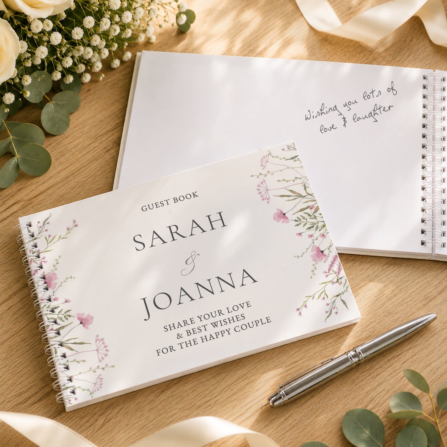 A personalised spiral-bound guest book with floral illustrations on the cover is placed on a wooden surface, alongside another open guest book with handwritten text, a silver pen, and eucalyptus leaves.