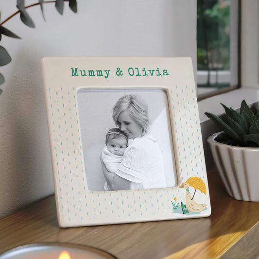 Personalised photo frame with a picture of a woman holding a baby, placed on a surface with plants in the background.