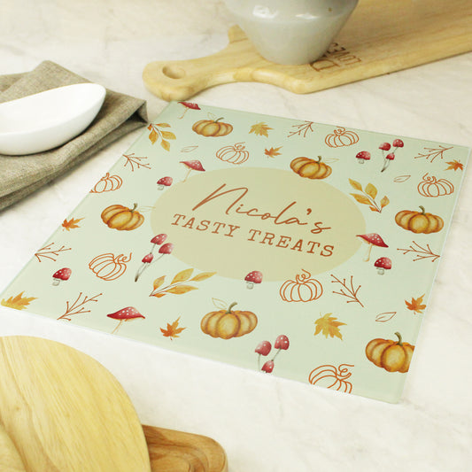 Personalised glass chopping board with autumn design and 'Nicola's Tasty Treats' text on a kitchen counter.