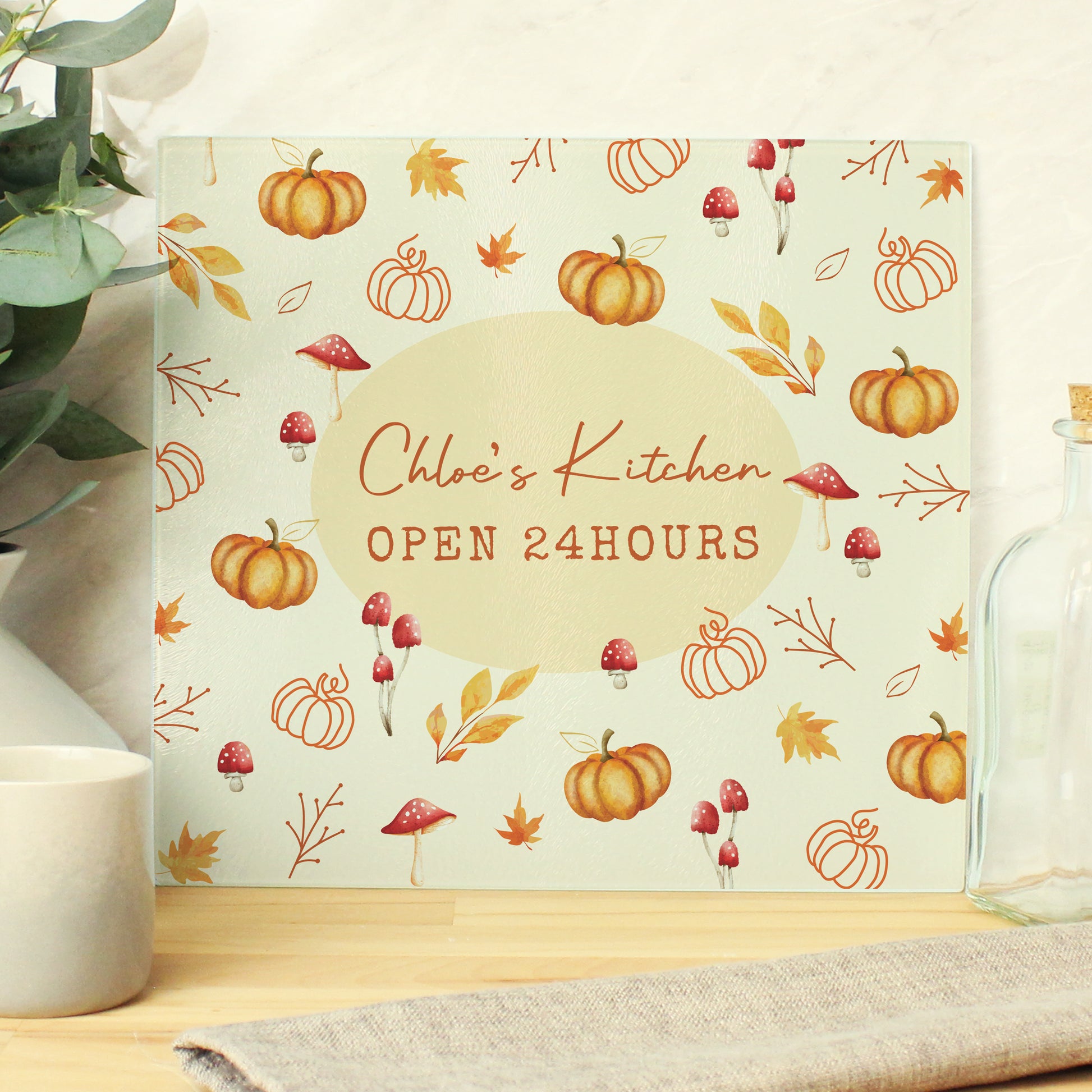 Personalised glass chopping board with autumn pumpkin design and 'Chloe's Kitchen Open 24 Hours' text on a wooden surface.