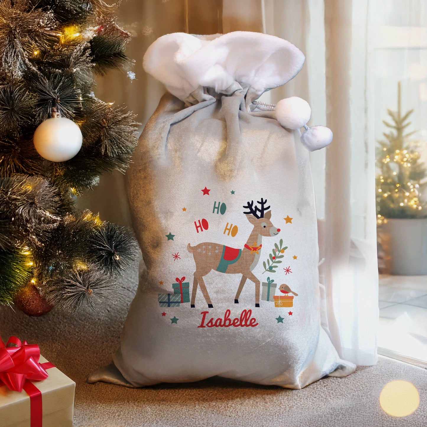 Decorative Christmas sack with reindeer design and 'Isabelle' text, next to a Christmas tree and gift box.