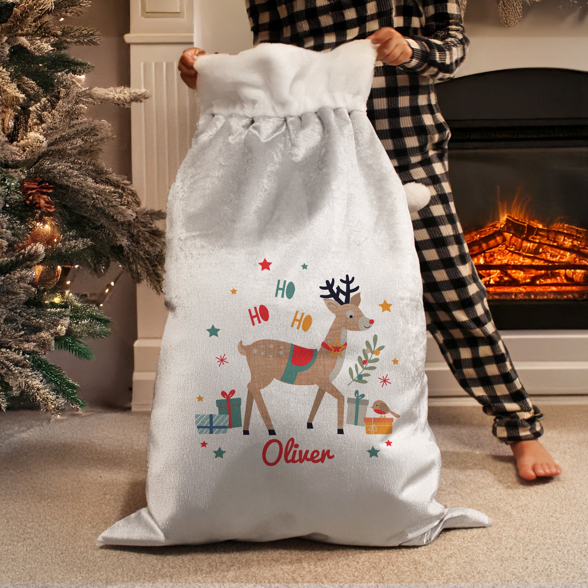Person holding a Christmas-themed sack with a reindeer design and 'Ho Ho Ho Oliver' text, in front of a fireplace.