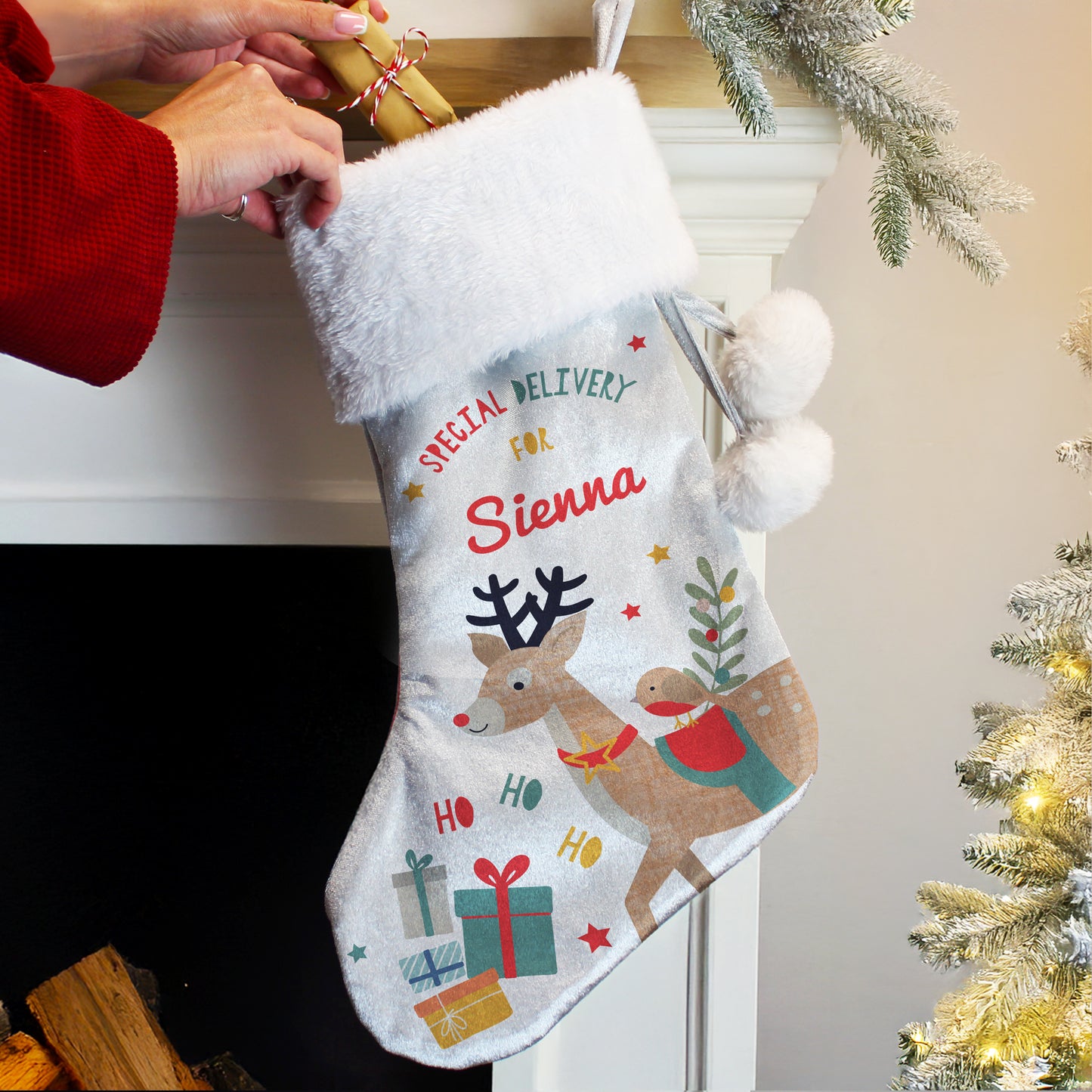 Personalised Christmas stocking with a reindeer design and text, hanging by a fireplace with a Christmas tree in the background.