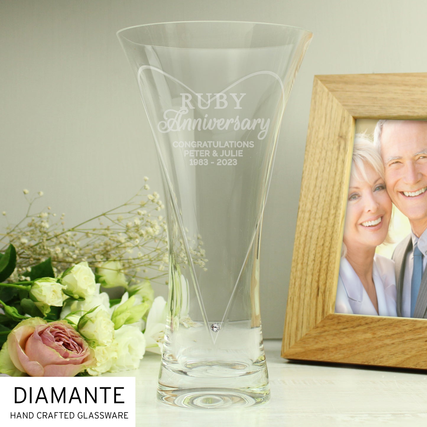 Clear glass vase with 'Ruby Anniversary' engraving next to a wooden photo frame with a couple's portrait.