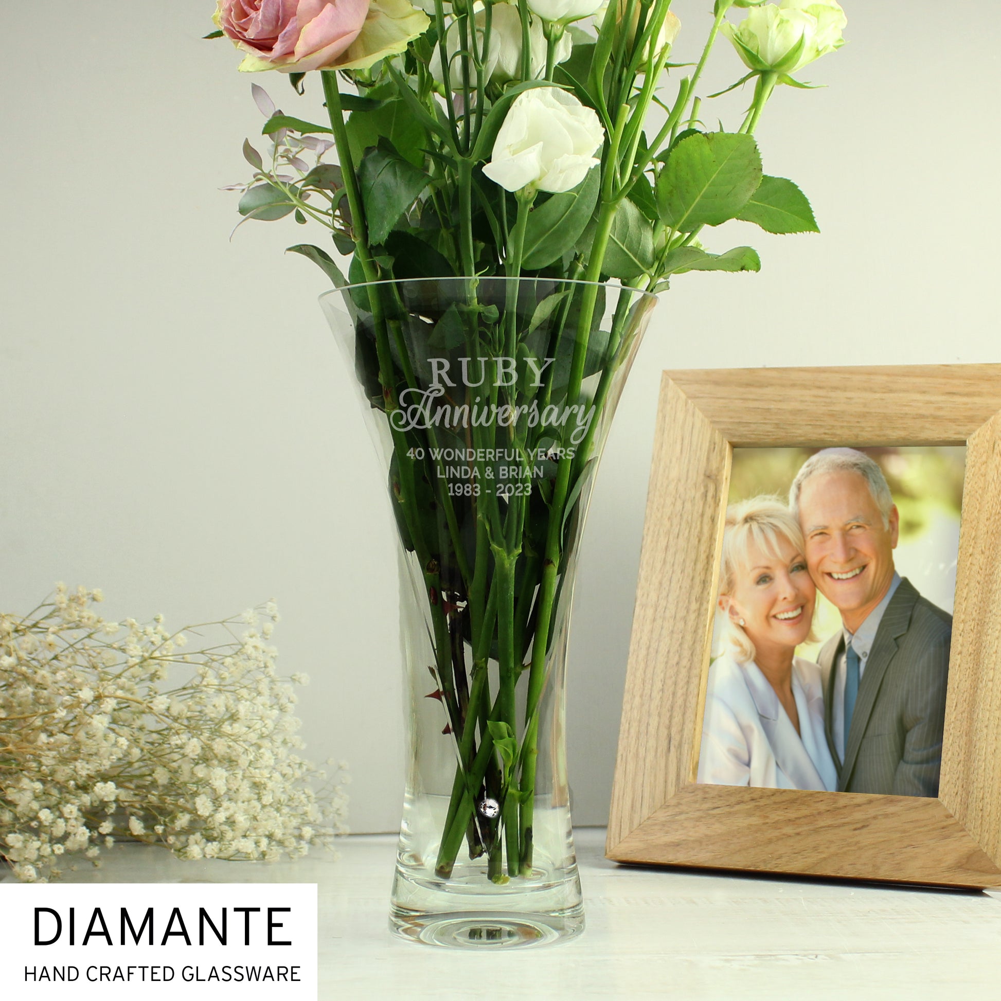 Clear glass vase with engraved 'Ruby Anniversary' text, filled with flowers, next to a wooden frame with a couple's photo.