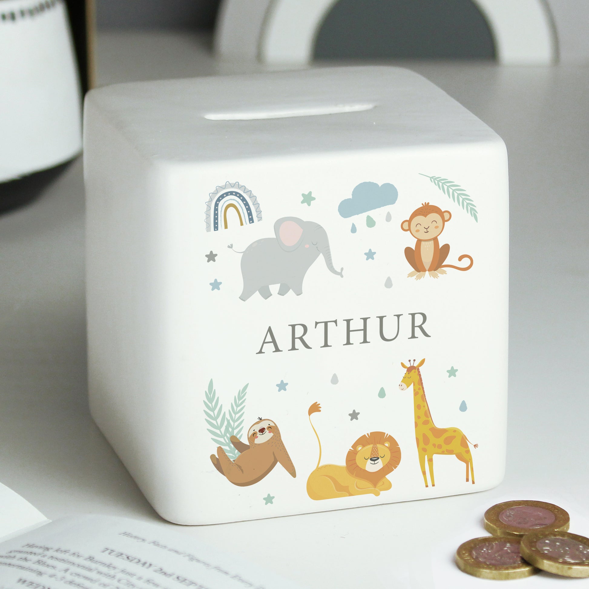 White ceramic square money box with safari animal and rainbow images  shown on a white shelf alongside open book and coins.