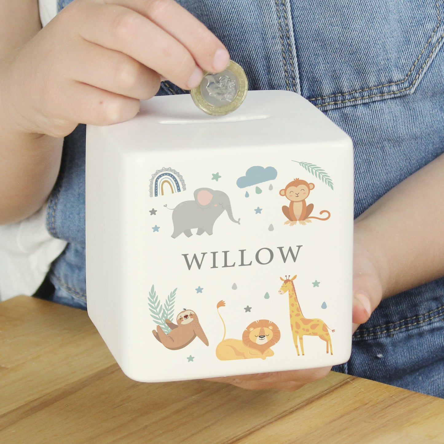 White ceramic square money box with the name 'WILLOW' with safari animal and rainbow images shown with someone holding a pound coin above it.