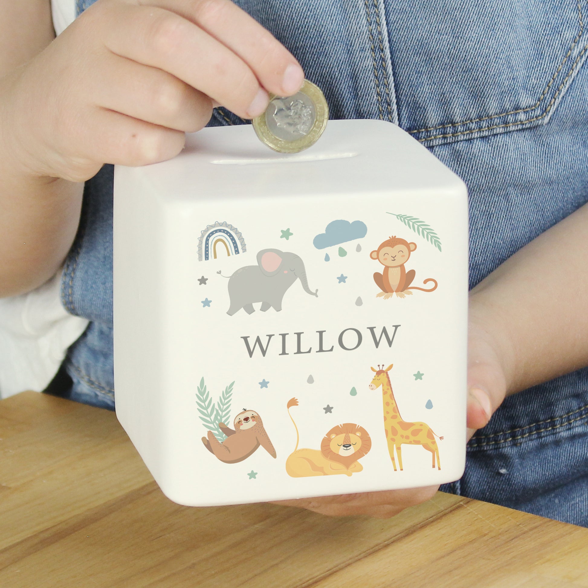White ceramic square money box with the name 'WILLOW' with safari animal and rainbow images shown with someone holding a pound coin above it.