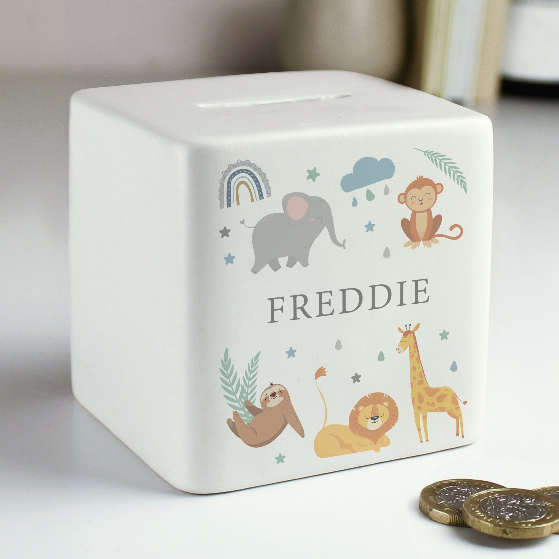 White ceramic square money box with the name 'FREDDIE' with safari animal and rainbow images  shown on a white shelf alongside coins.