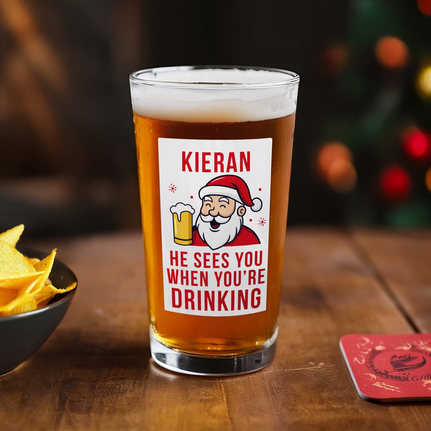 Beer glass with a Santa Claus label on a wooden table with chips and a coaster.