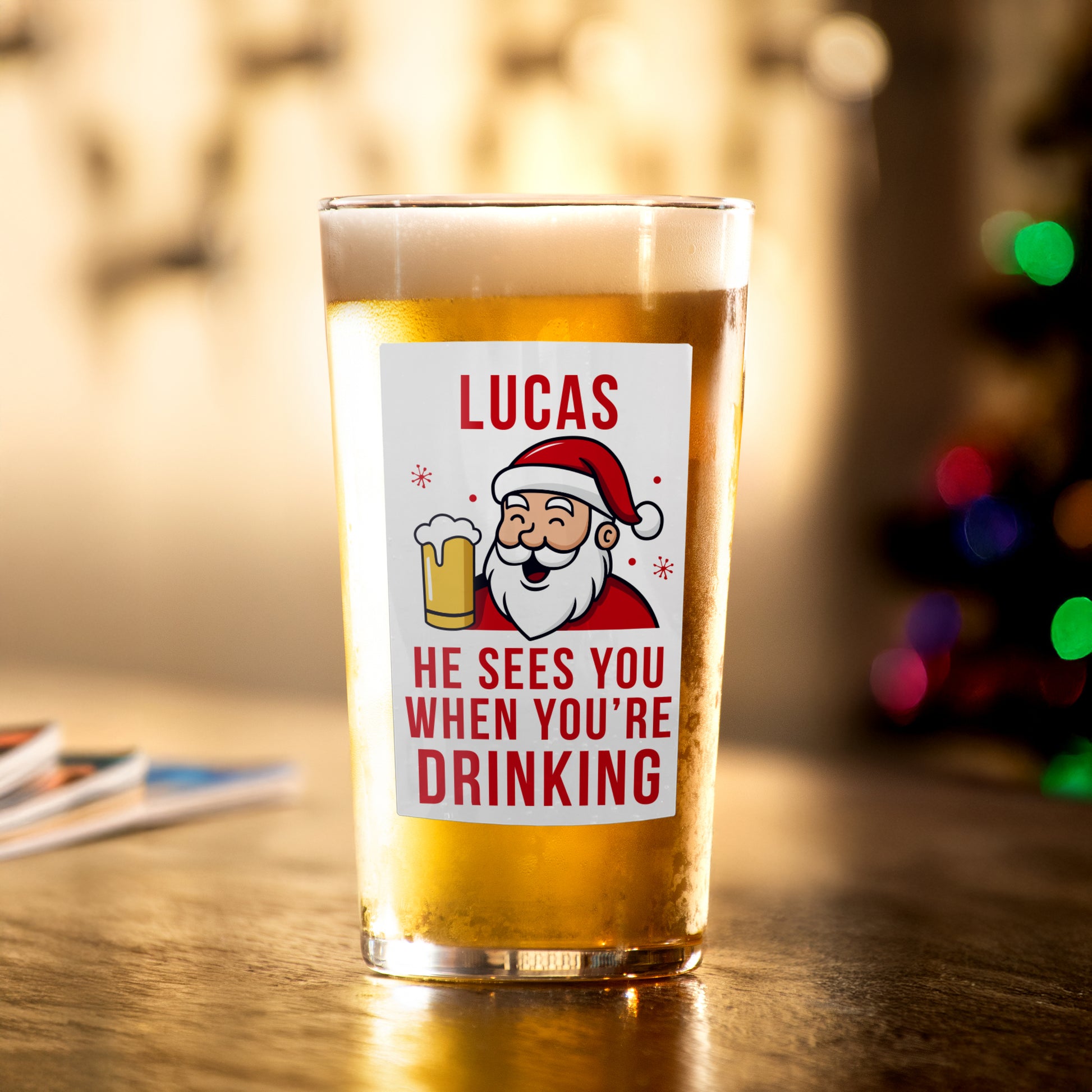 Beer glass with Santa Claus label and text 'Lucas He sees you when you're drinking' on a wooden table.