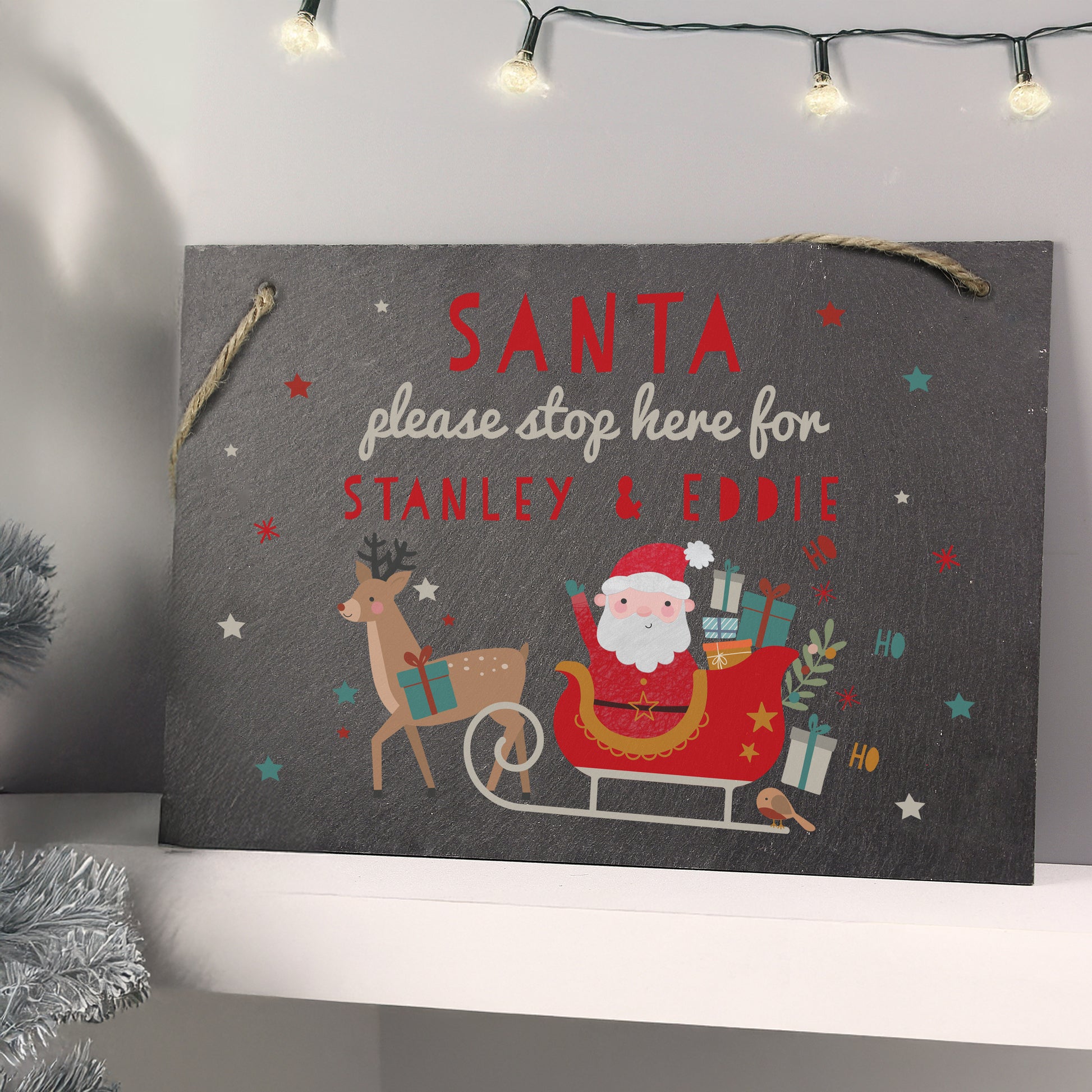 Slate Santa Stop Here sign shown standing on a shelf below Christmas lights. Text reads anta please stop here for Stanley & Eddie