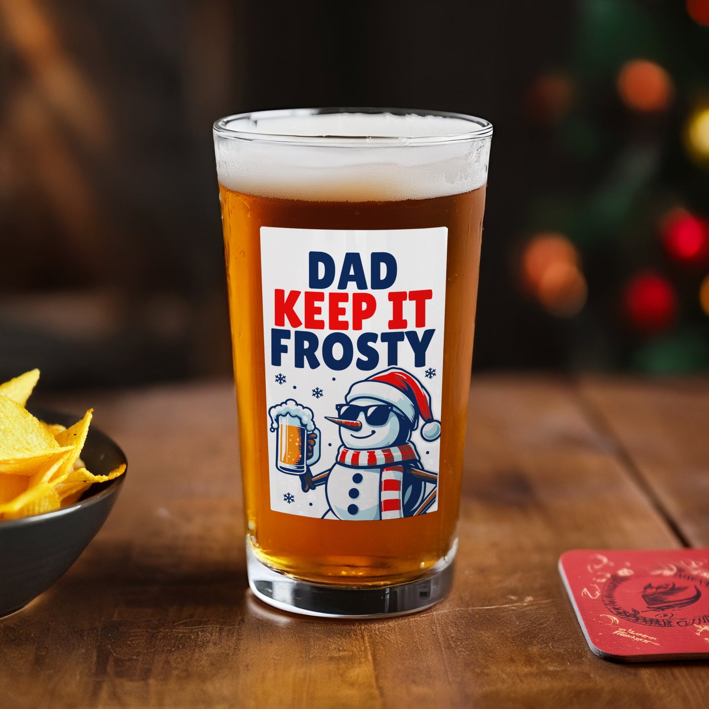 Beer glass with 'Dad Keep It Frosty' label on a wooden table with chips and a coaster.