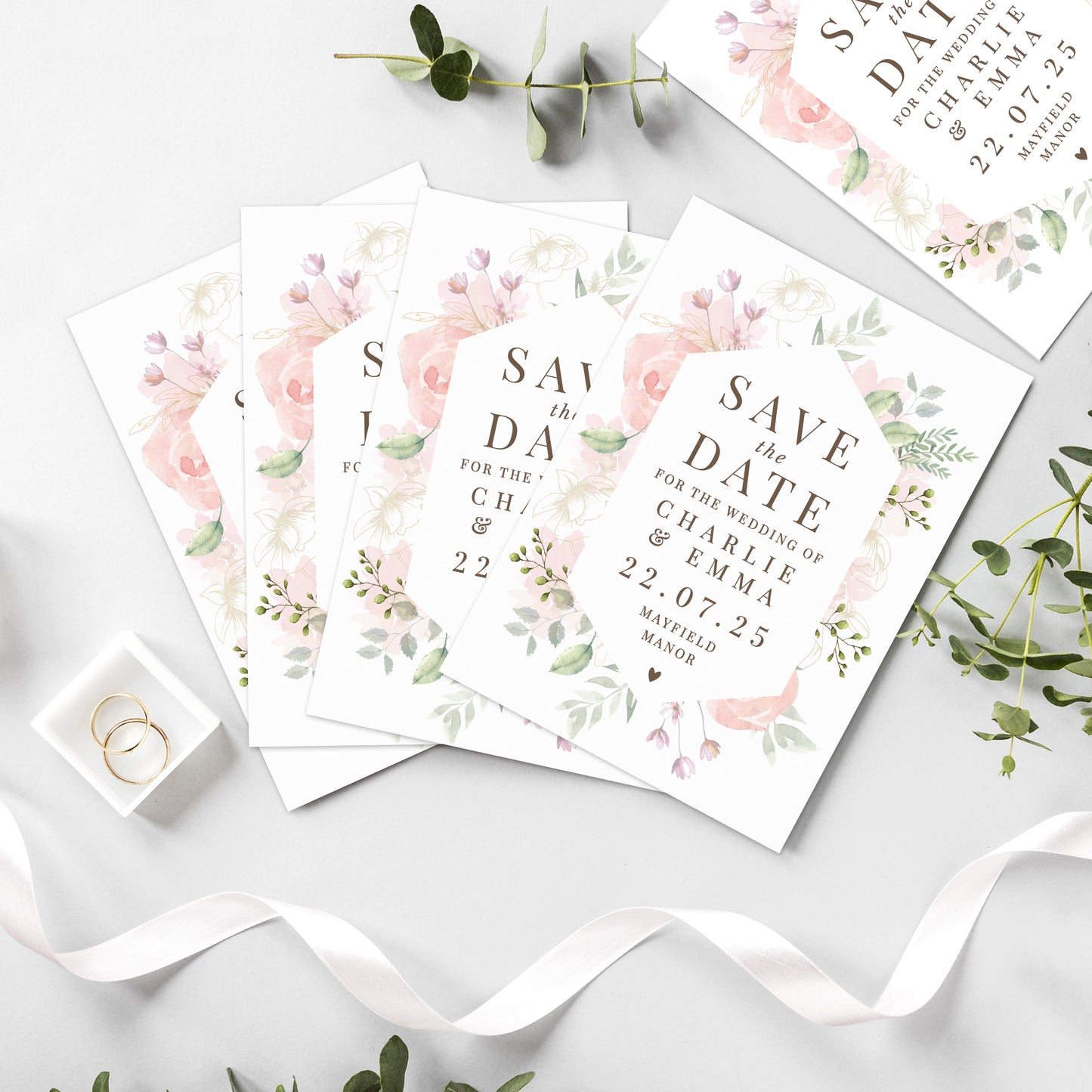 Floral 'Save the Date' cards on a white surface with greenery and a ring.