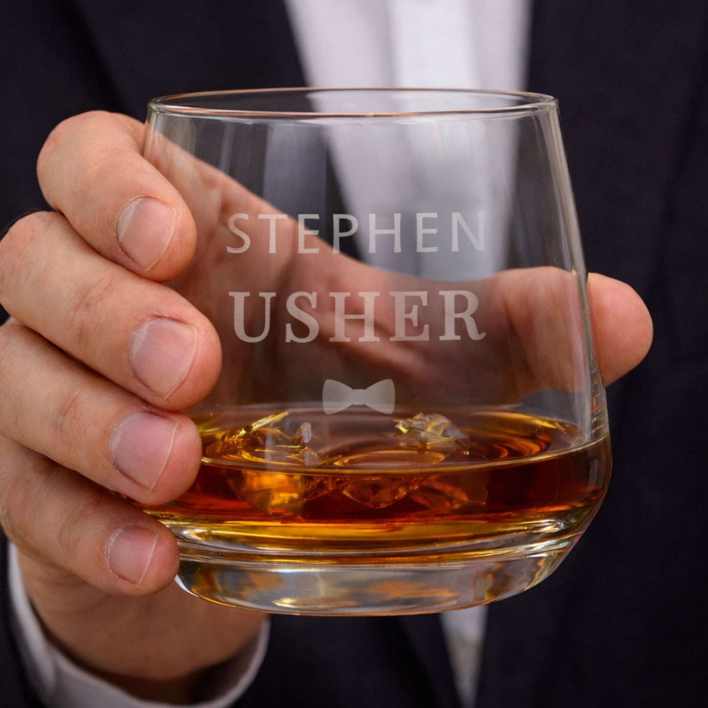 Hand holding a glass of whisky with 'Stephen Usher' engraved on it, against a blurred background.