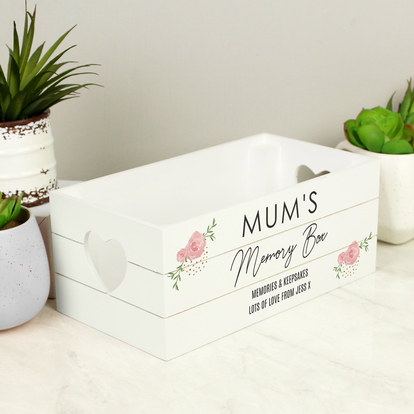 White wooden storage crate with pink rose design on the corners. Heart shaped cut out handles. Shown among plants.