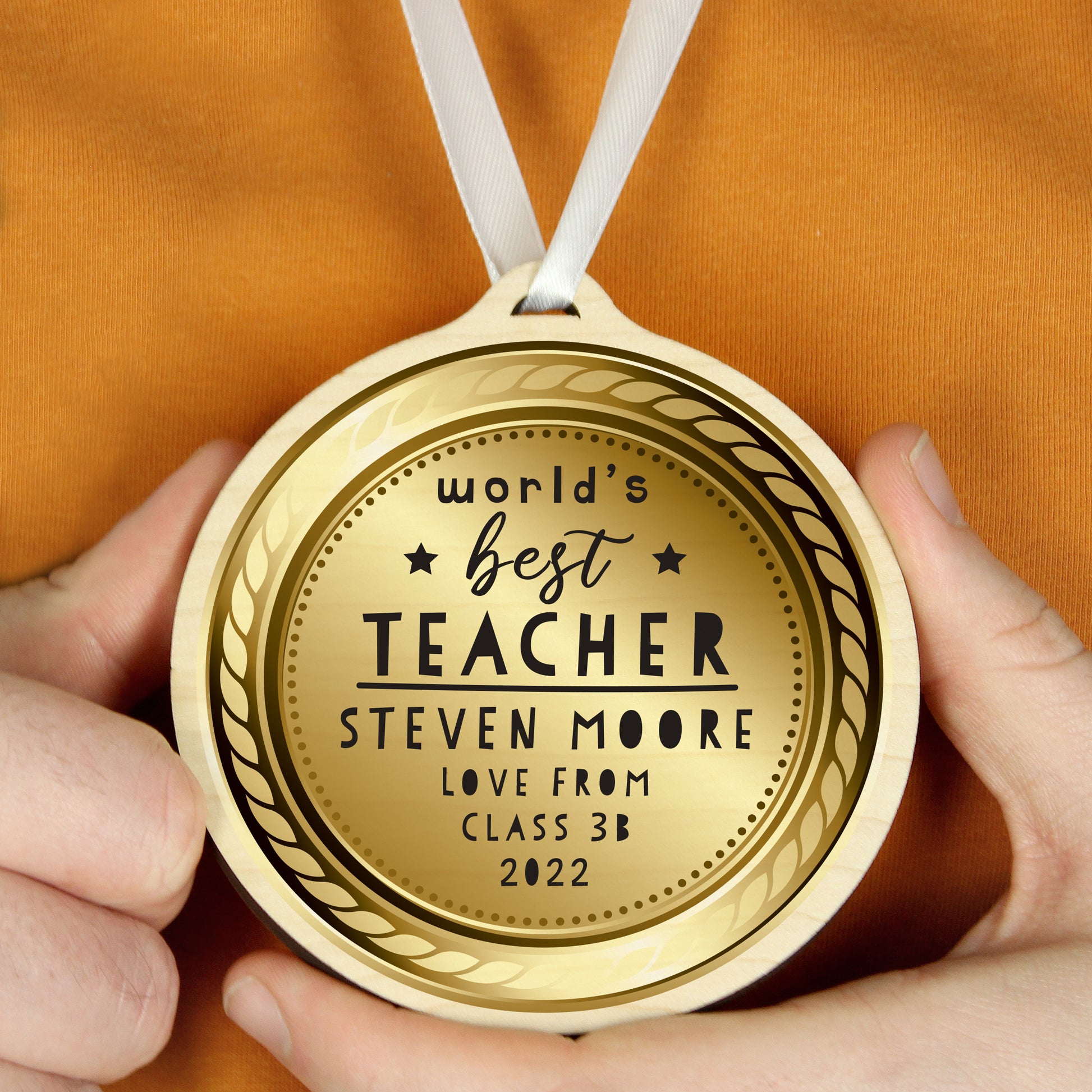 Gold coloured medal with 'World's Best Teacher' text held by hands against an orange background