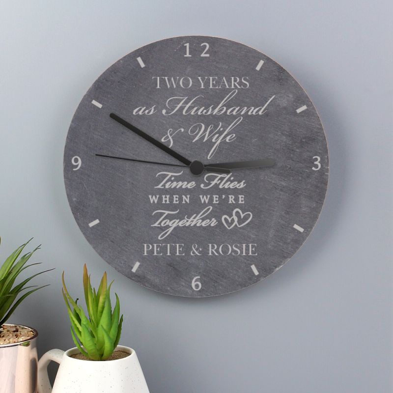 Wall clock with personalised message on a grey wall with plants below.